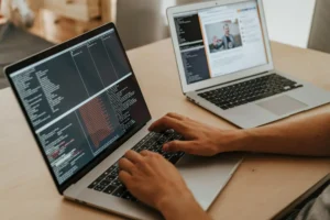 Imagem mostrando um desenvolvedor trabalhando em dois laptops, focando na criação de um sitemap e análise de código. A tela à esquerda exibe linhas de código, enquanto a direita mostra interação em uma plataforma online.