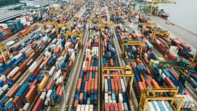 Imagem aérea de um grande porto de cargas com containers coloridos, guindastes e caminhões, ilustrando os desafios e soluções na cadeia de suprimentos industrial.